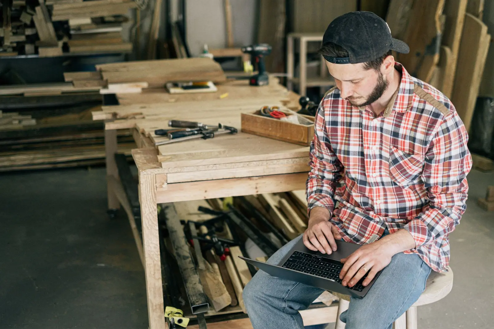 A carpenter in a workshop, working on a laptop on their lap